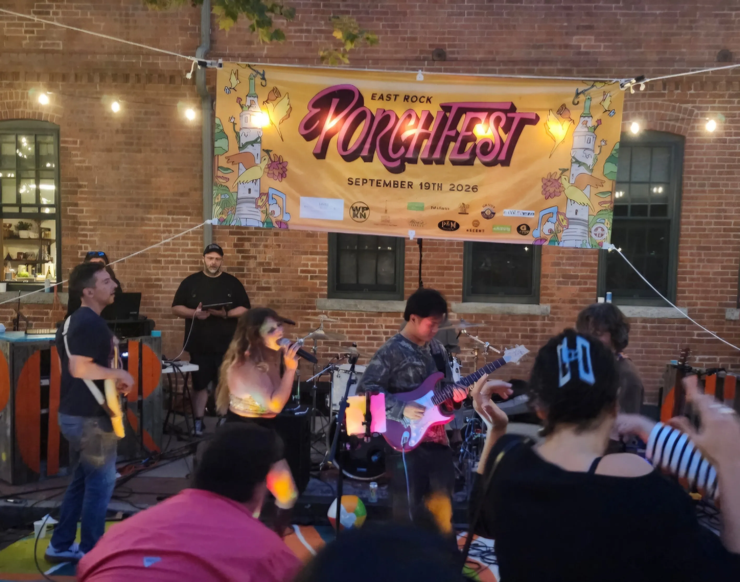 Neighbors gather outdoors at an East Rock community event, with a live band playing in front of a brick building under string lights.
